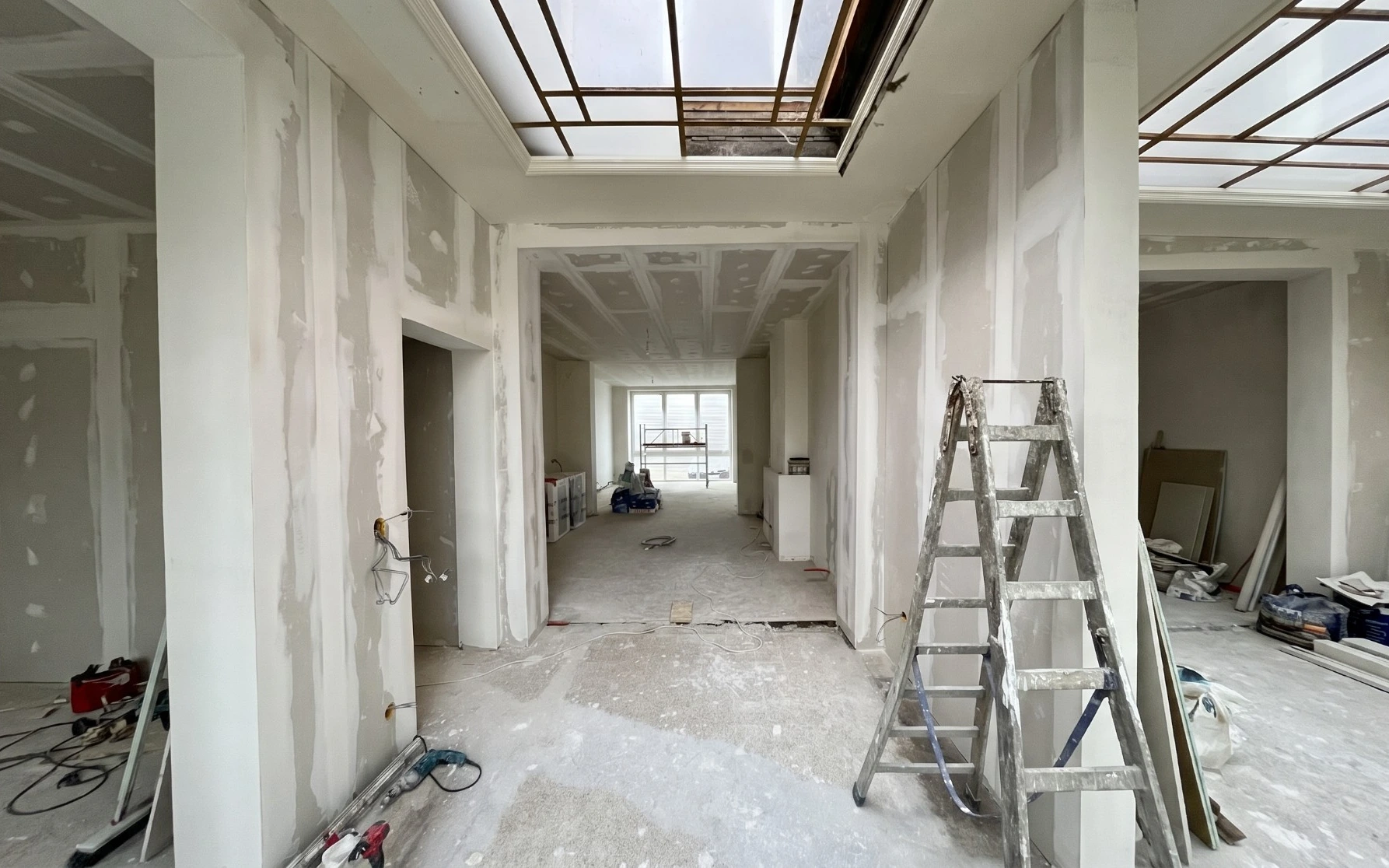 Hallway before renovation: taped plasterboard walls and ceilings, open ceiling frames, a stepladder and tools on a bare screed floor