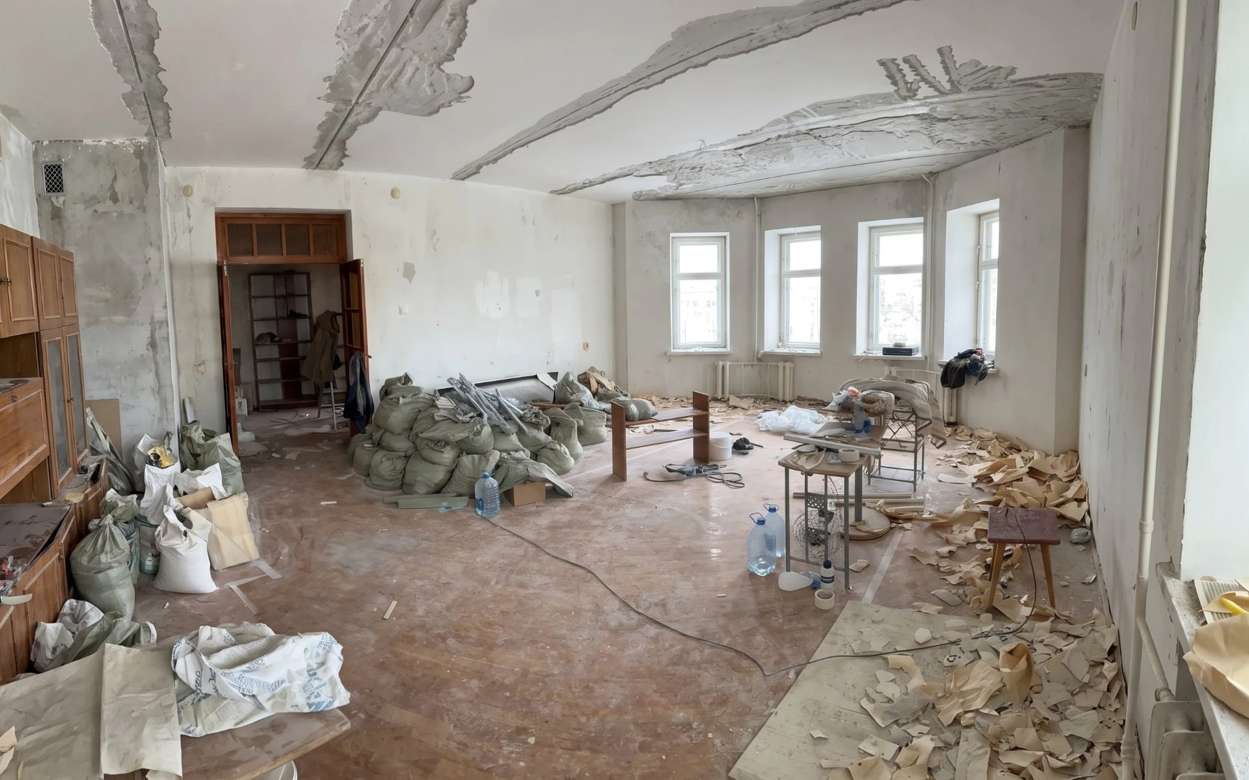 The same room before renovation: stripped ceiling and walls, old wooden wall units, construction bags and torn wallpaper on the floor